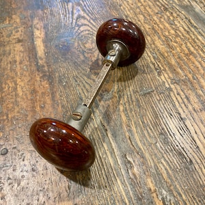 May include: A door knob set featuring two round, dark brown, marbled, translucent knobs connected by a square metal spindle. Each knob is attached to a silver-toned metal base with a visible screw, showcasing a classic design with a polished finish.