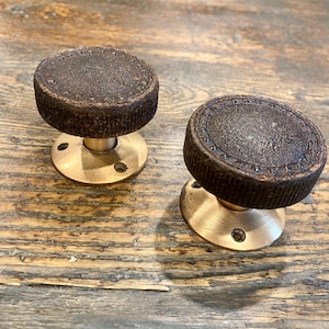 以下が含まれることがあります： 暗い質感の風化した茶色の金属製トップ、溝付きの縁、装飾的なドットの境界線が特徴の2つの円形キャビネットノブ。各ノブは、2つのネジ穴がある滑らかな真鍮色の円形ベースプレートに取り付けられています。