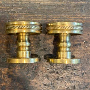 May include: Two antique brass door knobs. Each knob has a round, flat base, a decorative, hourglass-shaped stem, and a top with a rope-like design. The knobs are a warm, golden color and appear to be vintage.