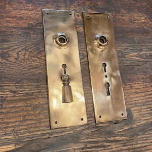 May include: Two antique brass door plates. Each plate features a keyhole, a decorative knob, and a circular opening for the door handle. The plates are rectangular with rounded edges and are mounted with small screws.