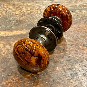 May include: A pair of round door knobs featuring a marbled brown and amber pattern with a glossy finish. The knobs are connected by a central metal spindle and include two circular dark metal mounting plates with visible screw holes for installation.