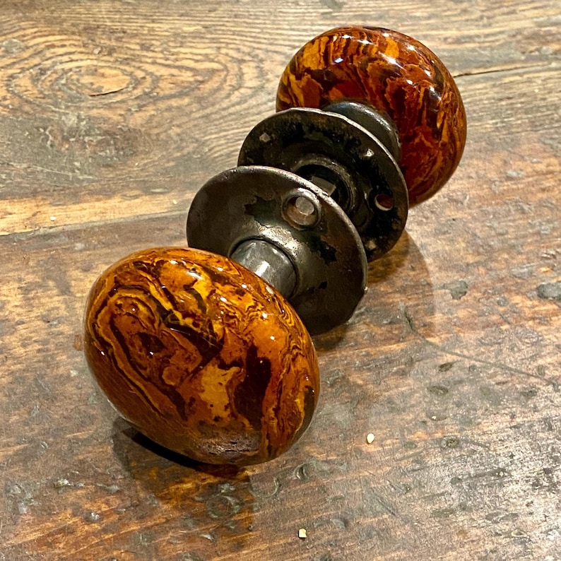 May include: A pair of round door knobs featuring a marbled brown and amber pattern with a glossy finish. The knobs are connected by a central metal spindle and include two circular dark metal mounting plates with visible screw holes for installation.