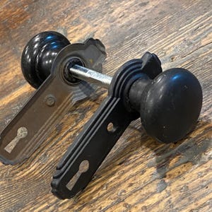 Può includere: Una coppia di pomelli neri per porta con placche rettangolari abbinate dotate di fori per serratura. I due pomelli sono collegati da un perno metallico centrale. La ferramenta presenta una finitura opaca e un design strutturato con bordi.