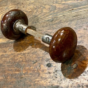 May include: A pair of round, dark brown, polished door knobs connected by a central metal square spindle. Each knob features a metallic collar with a visible screw, showcasing a glossy, mottled finish on the surface of the knobs.