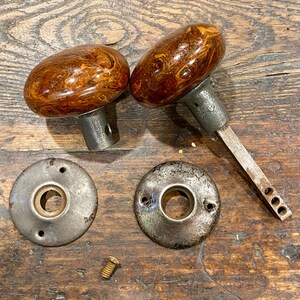 May include: Two oval-shaped door knobs with a polished brown and gold marbled finish, featuring grey metal bases. One knob includes a long square spindle. Two circular grey metal backplates with screw holes and one small brass screw are included.