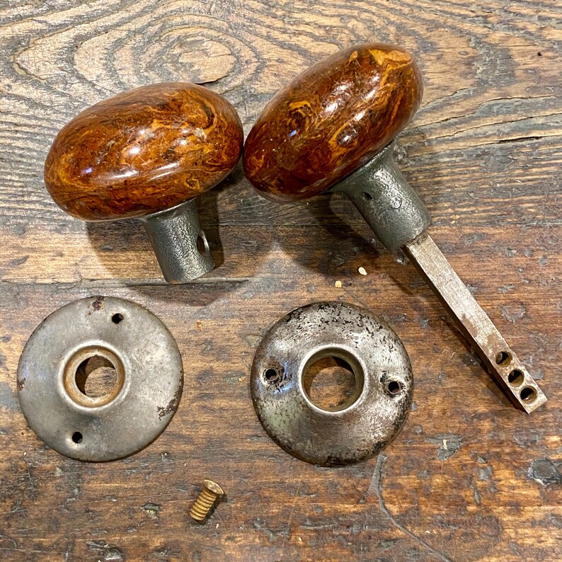 May include: Two oval-shaped door knobs with a polished brown and gold marbled finish, featuring grey metal bases. One knob includes a long square spindle. Two circular grey metal backplates with screw holes and one small brass screw are included.
