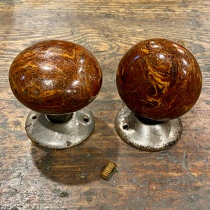 May include: Two round door knobs with a glossy brown and tan marbled pattern. Each knob is attached to a circular, weathered silver-toned metal base with two screw holes. A single small brass-colored screw is positioned between the two knobs.