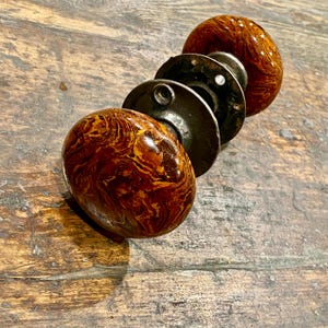 May include: A pair of round door knobs featuring a glossy, marbled finish in shades of dark brown, amber, and gold. The knobs are connected by a central metal spindle and include circular dark metal mounting plates with visible screw holes.
