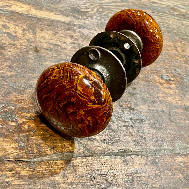 May include: A pair of round door knobs featuring a glossy, marbled finish in shades of dark brown, amber, and gold. The knobs are connected by a central metal spindle and include circular dark metal mounting plates with visible screw holes.