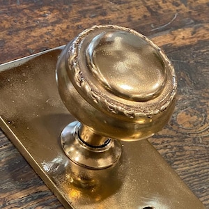 May include: A close-up of a vintage brass door knob and plate. The knob is round with a decorative edge, and the plate is rectangular. The brass has a warm, golden hue, and the design suggests an antique style.