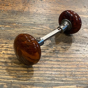 May include: A door knob set featuring two round, polished, dark brown and amber marbled knobs connected by a central metal square spindle. Each knob is attached to a silver-toned metal base with a visible screw, showcasing a smooth, glossy finish.
