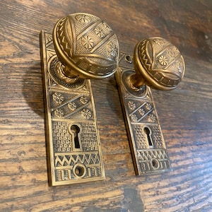 Puede incluir: Dos pomos de puerta de latón antiguo con placas rectangulares a juego. Cada pomo tiene una forma esférica con intrincados grabados geométricos y florales. Las placas rectangulares muestran un patrón a juego, una cerradura central y orificios para tornillos.