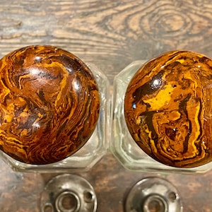May include: Two round cabinet knobs featuring a glossy, marbled pattern in shades of amber, dark brown, and gold. Each knob is attached to a clear, faceted glass base and includes a circular metal mounting plate with two screw holes at the bottom.