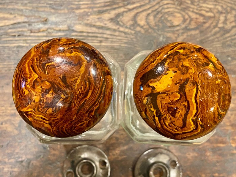 May include: Two round cabinet knobs featuring a glossy, marbled pattern in shades of amber, dark brown, and gold. Each knob is attached to a clear, faceted glass base and includes a circular metal mounting plate with two screw holes at the bottom.
