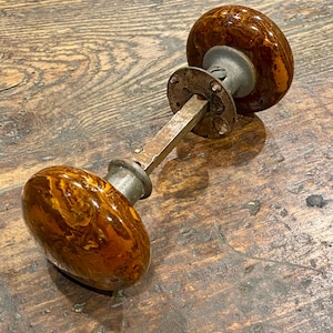 May include: A door knob set featuring two round, glossy, brown and amber marbled knobs connected by a square metal spindle. The assembly includes a circular metal mounting plate with visible screw holes and a weathered, metallic finish on the hardware.