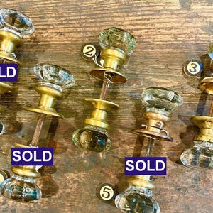 May include: Five sets of door knobs featuring clear, faceted glass handles attached to brass-toned metal hardware. Each set includes a central spindle and circular mounting plates. The items are numbered one through five, with three marked SOLD.