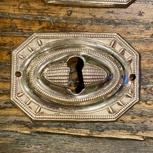 May include: A metallic, rectangular escutcheon plate with clipped corners, featuring a central keyhole opening. The surface displays an embossed, decorative border with repeating linear patterns and a textured, oval-shaped center detail.