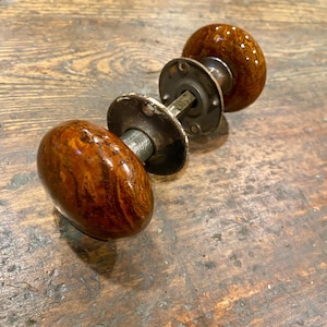 May include: A pair of round, brown, marbled-pattern door knobs with a glossy finish. The knobs are connected by a central metal spindle and feature circular, dark-colored metal mounting plates with visible screw holes for installation.