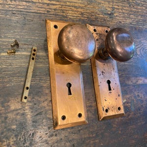 Puede incluir: Dos manijas de puerta antiguas de cobre con placas traseras rectangulares a juego. Los pomos son redondos y tienen un acabado deslustrado. Las placas traseras tienen cerraduras y orificios para tornillos. También hay una pequeña placa y tornillos de latón.