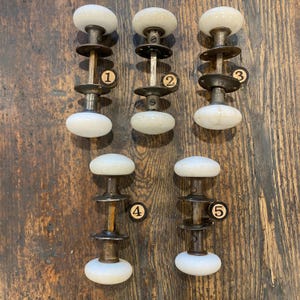 May include: Five antique door knobs are displayed. Each has a white, oval porcelain handle, connected by a metal rod and plates. The metal parts show signs of age and wear, enhancing their vintage charm. The knobs are arranged on a wooden surface.