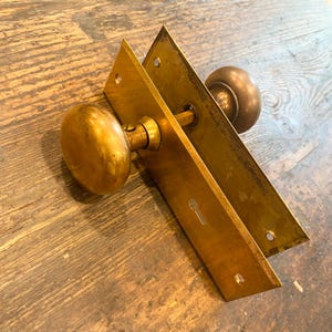 May include: Vintage brass door knob set featuring a keyhole and two rectangular plates. The door knob is spherical and has a warm, golden tone. The plates are rectangular with rounded edges. The set is designed for a door and is made of brass.