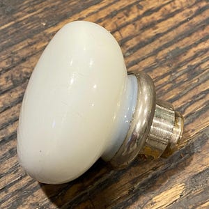 May include: A white, oval-shaped cabinet knob with a silver metal base and a brass screw. The knob is set against a rustic, wooden surface.