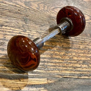 May include: A door knob set featuring two round, polished, dark brown and reddish-marbled knobs connected by a central square metal spindle. The knobs have a smooth, glossy finish with a swirling pattern, attached to dark metallic circular bases.