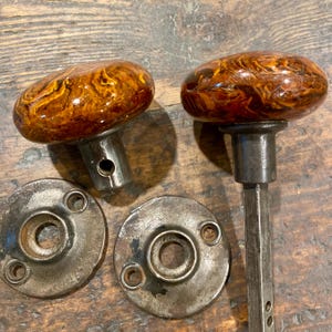 May include: Two round door knobs with a glossy, marbled amber and brown finish, each featuring a metallic base. The set includes two circular metal backplates with screw holes, and one knob is attached to a square metal spindle shaft.