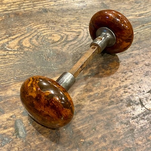 May include: A door knob set featuring two round, polished brown and amber marbled knobs with a glossy finish. The knobs are connected by a square metal spindle and include silver-toned metal collars at the base of each knob, showing a visible screw.