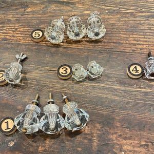 May include: A collection of clear glass cabinet knobs with faceted designs, arranged on a wooden surface. Each knob has a metal screw and is paired with a small, round wooden disc with a number from 1 to 5.