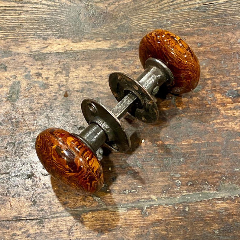 May include: A set of two round, marbled brown and amber glass door knobs connected by a central metal spindle and two circular metal mounting plates with visible screw holes, featuring a polished finish on the glass and a dark, weathered metal.