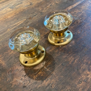 May include: Two vintage glass door knobs with brass bases. The clear glass knobs have a starburst design. The brass bases have a circular shape with four screw holes. The knobs are set against a dark wood surface.