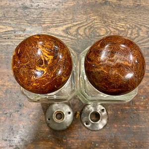 May include: Two clear glass door knobs with spherical, polished brown and gold marbled patterns. Each knob is paired with a circular, weathered gray metal mounting plate featuring a central hole and small screw openings for installation.