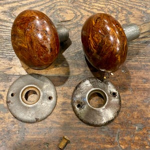 May include: Two oval-shaped door knobs with a glossy, marbled brown and amber finish, each attached to a metal spindle. Included are two circular, weathered metal backplates with screw holes and one small, threaded metal screw for installation.