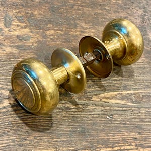 Può includere: Due maniglie per porte antiche in ottone con un classico design rotondo. Le maniglie sono collegate da un'asta metallica e sono appoggiate su una superficie di legno rustico. L'ottone presenta una calda patina invecchiata.