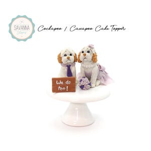 May include: A white cake topper featuring two small, white dogs dressed in wedding attire. The groom dog wears a purple tie and the bride dog wears a lavender dress and a pearl necklace. The dogs are holding a sign that says "We do too!"