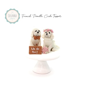 May include: White ceramic cake topper featuring a bride and groom French poodle. The bride poodle wears a white dress and a pink flower crown. The groom poodle wears a brown bow tie. The couple stands on a white cake stand with a sign that reads "We do too!"