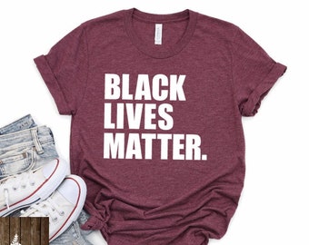 May include: A maroon t-shirt with the text "Black Lives Matter" printed in white on the front. The shirt is photographed on a white background with a pair of jeans and white sneakers.
