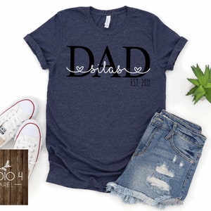 May include: A navy blue t-shirt with the text "DAD Silas EST. 2021" printed on it. The text is in black with a white heart around the name Silas.