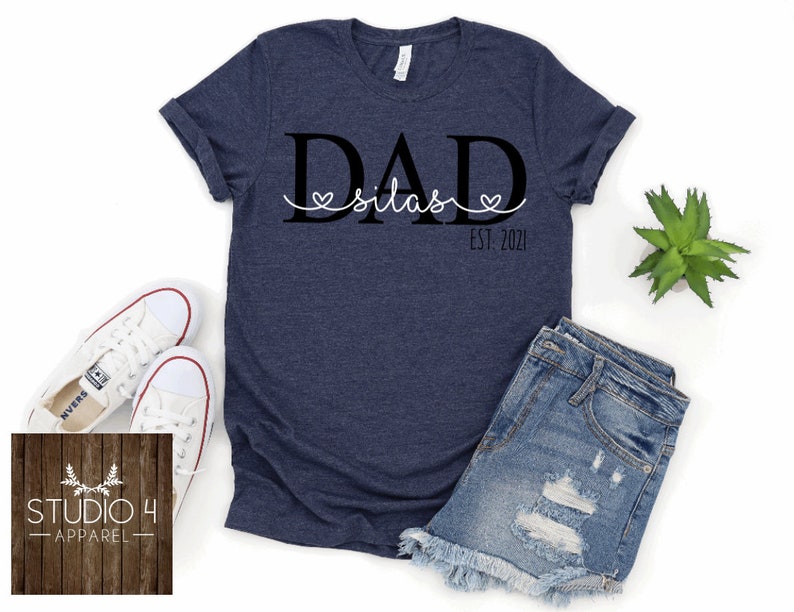 May include: A navy blue t-shirt with the text "DAD Silas EST. 2021" printed on it. The text is in black with a white heart around the name Silas.