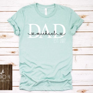 May include: A light blue t-shirt with the word "DAD" in white letters and the text "michael" in black script with hearts on either side. The text "EST. 2017" is printed below the word "DAD".