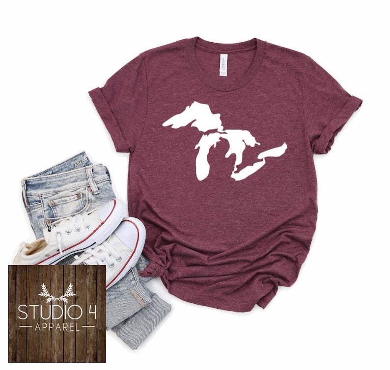 May include: A maroon t-shirt featuring a white outline of the Great Lakes. The shirt is laid flat on a white background with a pair of jeans and white trainers.