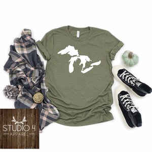 May include: A green t-shirt with a white outline of the Great Lakes on the front. The shirt is lying flat on a white background with other items around it, including a plaid scarf, a small pinecone, a woven ball, a small pumpkin, and two black and white high-top trainers.