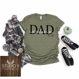 May include: Green t-shirt with black text that reads "DAD Silas EST. 2021" with two small hearts on either side of the name.