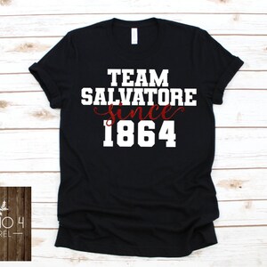 May include: Black t-shirt with white text that reads "Team Salvatore since 1864". The word "since" is written in cursive and the year 1864 is in red.