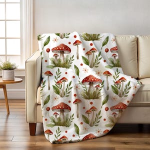 May include: A white throw blanket draped over a beige sofa. The blanket features a repeating pattern of red and white spotted mushrooms, green leaves, and small white flowers. The blanket is made of a soft, plush material.