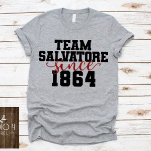 May include: Gray t-shirt with black text that reads "Team Salvatore since 1864". The word "since" is in red cursive.