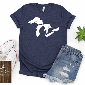 May include: A navy blue t-shirt featuring a white outline of the Great Lakes. The shirt is displayed on a white background with a pair of white trainers, a pair of blue denim shorts, and a small green plant.