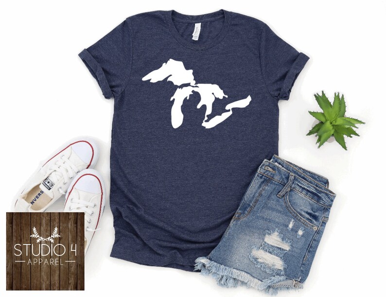May include: A navy blue t-shirt featuring a white outline of the Great Lakes. The shirt is displayed on a white background with a pair of white trainers, a pair of blue denim shorts, and a small green plant.