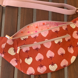 May include: A rust-colored fanny pack with a strawberry pattern. The bag has two zippered compartments on the front and a pink strap. The interior lining is a pink fabric with a polka dot pattern. The zippers are a rose gold color.